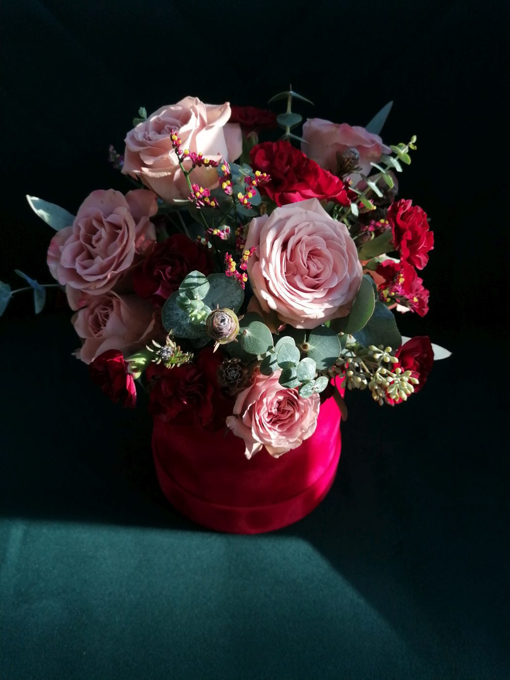 The soft shade of beige roses contrasts with the red of the carnations and the box