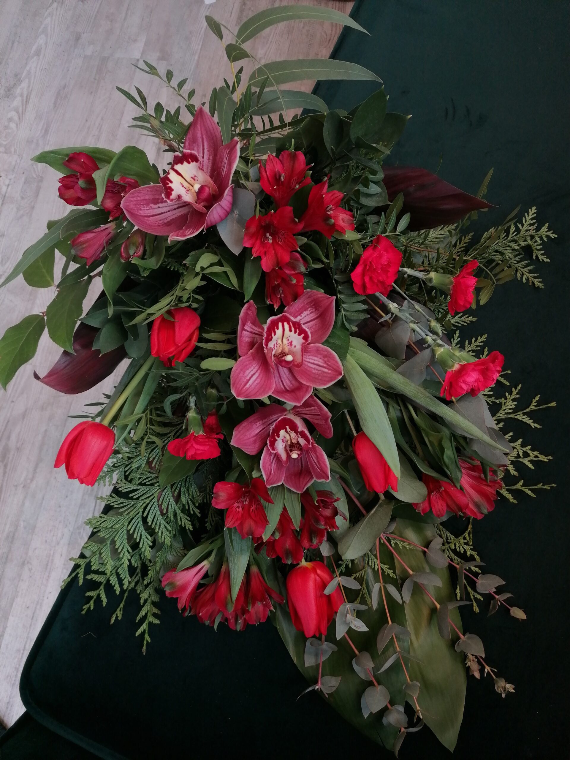 Wiązanka w czerwieni to elegancki bukiet pożegnalny, zarówno dla kobiety jak i mężczyzny. Składa się z storczyków, tulipanów, alstremerii i goździków w otoczeniu różnorodnej zieleni.