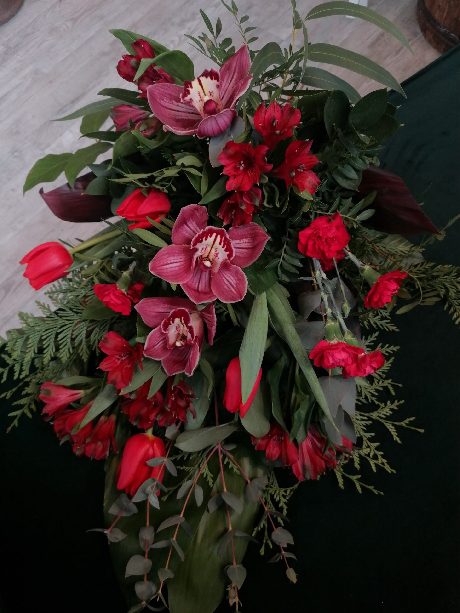 Букет в червоній гамі - це елегантний прощальний букет для чоловіків і жінок. Він складається з орхідей, тюльпанів, альстромерій та гвоздик в оточенні різноманітної зелені.