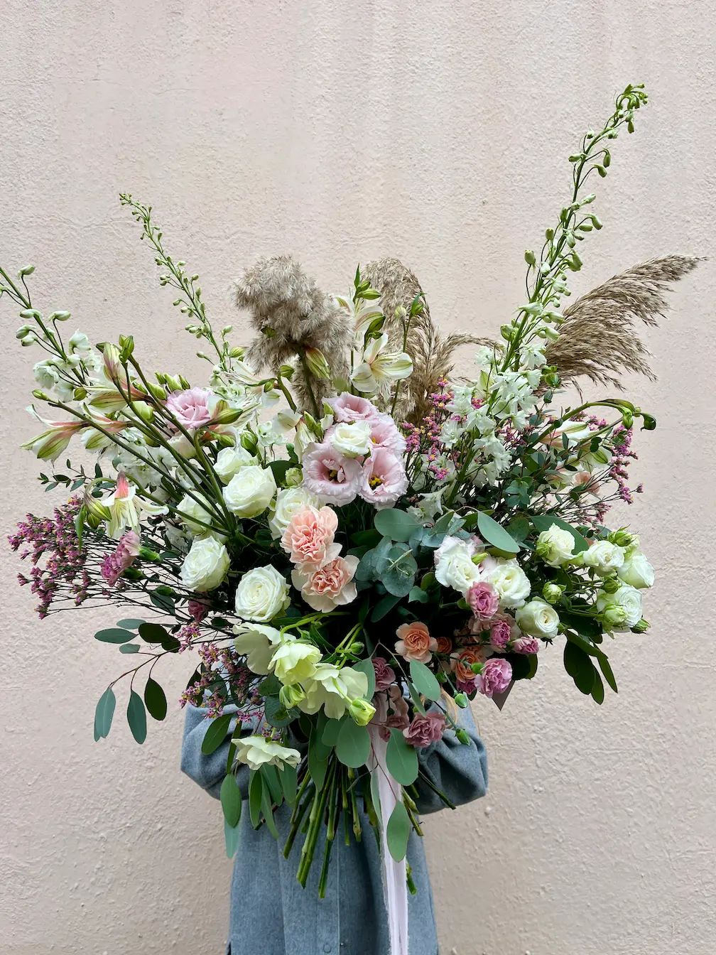 Osoba trzyma duży, naturalny bukiet z jasnych róż, eustomy, zieleni i traw ozdobnych
